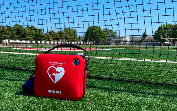 A Life Saving AED Defibrillator On A Turf Field With A Net Behind It For Safety Of Athletes, Coaches And Fans.